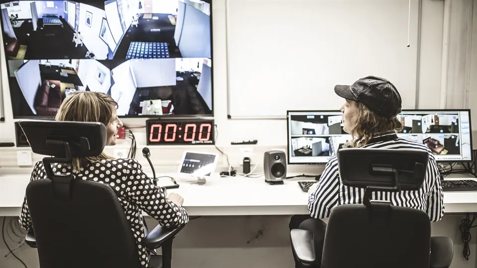 Two people are sitting at a long work table. In front of them are several computer screens, a microphone and other technical equipment.